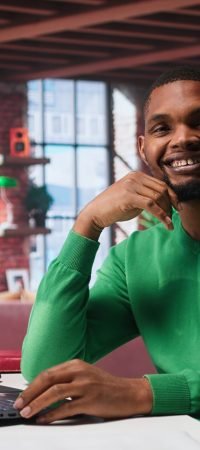African american remote worker solving his daily freelancing tasks on laptop, working in relaxation from the comfort of his modern loft. Tech savvy black man internet user, multitasking. Camera B.