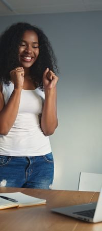Human facial expressions and emotions. Euphoric emotional young mixed race female clenching fists, being excited with good news, success or promotion at work, standing at desk with laptop and diary
